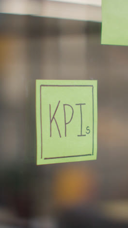 Vertical-Video-Close-Up-Of-Woman-Putting-Sticky-Note-With-KPIs-Written-On-It-Onto-Transparent-Screen-In-Office-